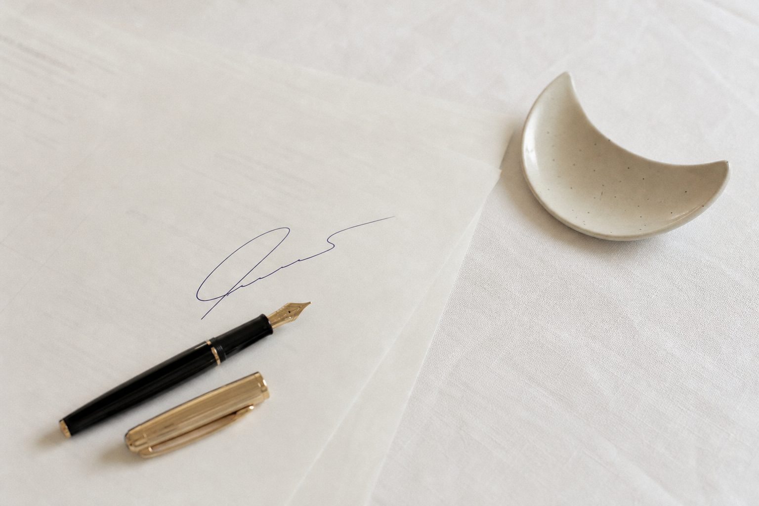 A paper contract and pen on white linen beside a small crescent ceramic dish